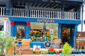 A charming antique shop display featuring a variety of vintage items. The exterior is painted dark blue with the word 'ANTIQUES' prominently displayed above the entrance. The storefront is filled with various antique objects, including wooden crates, earthen pots, a ship's wheel, and a stack of vintage suitcases. Red fire buckets hang on either side of the entrance. The interior is bustling with a diverse collection of antiques, adding to the shop's allure.