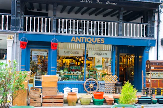 A charming antique shop display featuring a variety of vintage items. The exterior is painted dark blue with the word 'ANTIQUES' prominently displayed above the entrance. The storefront is filled with various antique objects, including wooden crates, earthen pots, a ship's wheel, and a stack of vintage suitcases. Red fire buckets hang on either side of the entrance. The interior is bustling with a diverse collection of antiques, adding to the shop's allure.