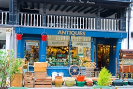 A charming antique shop display featuring a variety of vintage items. The exterior is painted dark blue with the word 'ANTIQUES' prominently displayed above the entrance. The storefront is filled with various antique objects, including wooden crates, earthen pots, a ship's wheel, and a stack of vintage suitcases. Red fire buckets hang on either side of the entrance. The interior is bustling with a diverse collection of antiques, adding to the shop's allure.