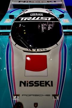 A top-down view of a racing car with the words 'Courage Competition' and 'Trust' on the windshield. The vehicle features a Nisseki logo and a large red and white emblem on the front. The color scheme includes light blue, pink, and white, with sleek and aerodynamic design elements. The Porsche logo is also visible on the vehicle.