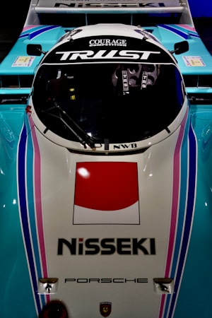 A top-down view of a racing car with the words 'Courage Competition' and 'Trust' on the windshield. The vehicle features a Nisseki logo and a large red and white emblem on the front. The color scheme includes light blue, pink, and white, with sleek and aerodynamic design elements. The Porsche logo is also visible on the vehicle.