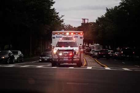 An ambulance with flashing lights is driving down a road, surrounded by cars on both sides. The scene takes place during dusk or dawn, as the sky is dimly lit. Trees line the road, and the atmosphere suggests urgency and movement.