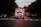 An ambulance with flashing lights is driving down a road, surrounded by cars on both sides. The scene takes place during dusk or dawn, as the sky is dimly lit. Trees line the road, and the atmosphere suggests urgency and movement.