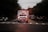 An ambulance with flashing lights is driving down a road, surrounded by cars on both sides. The scene takes place during dusk or dawn, as the sky is dimly lit. Trees line the road, and the atmosphere suggests urgency and movement.