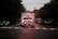 An ambulance with flashing lights is driving down a road, surrounded by cars on both sides. The scene takes place during dusk or dawn, as the sky is dimly lit. Trees line the road, and the atmosphere suggests urgency and movement.