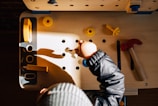 A child is playing with a wooden workbench toy set, featuring various plastic tools and parts such as a red hammer, yellow screws, and bolts. The child is wearing a striped hat and a dark jacket. Sunlight casts dynamic shadows on the workbench, creating a warm, playful environment.