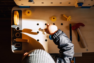 A child is playing with a wooden workbench toy set, featuring various plastic tools and parts such as a red hammer, yellow screws, and bolts. The child is wearing a striped hat and a dark jacket. Sunlight casts dynamic shadows on the workbench, creating a warm, playful environment.