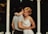 A couple embraces under a white pavilion with ornate columns. The woman, wearing a white dress, has short blond hair and appears at ease with her eyes closed. The man wears a gray shirt and has his arms around her. The background is dark, suggesting a serene evening setting.