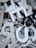 A jumble of variously sized and shaped metallic and plastic letters, predominantly in shades of silver, white, and black. Some letters are stacked on top of others, creating a chaotic and dense arrangement. A metallic screw is visible amidst the letters, adding to the industrial feel.