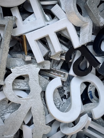 A jumble of variously sized and shaped metallic and plastic letters, predominantly in shades of silver, white, and black. Some letters are stacked on top of others, creating a chaotic and dense arrangement. A metallic screw is visible amidst the letters, adding to the industrial feel.