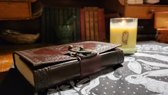 A leather-bound journal with an ornate metal clasp rests on a table covered with a black and white patterned cloth. A lit candle with an animal illustration and a warm glow adds a cozy ambiance. Books and wooden shelves create a rustic backdrop.