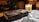 A leather-bound journal with an ornate metal clasp rests on a table covered with a black and white patterned cloth. A lit candle with an animal illustration and a warm glow adds a cozy ambiance. Books and wooden shelves create a rustic backdrop.