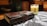 A leather-bound journal with an ornate metal clasp rests on a table covered with a black and white patterned cloth. A lit candle with an animal illustration and a warm glow adds a cozy ambiance. Books and wooden shelves create a rustic backdrop.
