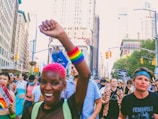 A diverse group of people is seen participating in a vibrant street parade in a city with tall buildings. The atmosphere is lively, featuring individuals expressing support for LGBTQ+ rights, as indicated by rainbow accessories and banners. One person with pink hair and a rainbow wristband is prominently raising a fist, while others display various messages on their clothing and signs.