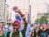 A diverse group of people is seen participating in a vibrant street parade in a city with tall buildings. The atmosphere is lively, featuring individuals expressing support for LGBTQ+ rights, as indicated by rainbow accessories and banners. One person with pink hair and a rainbow wristband is prominently raising a fist, while others display various messages on their clothing and signs.