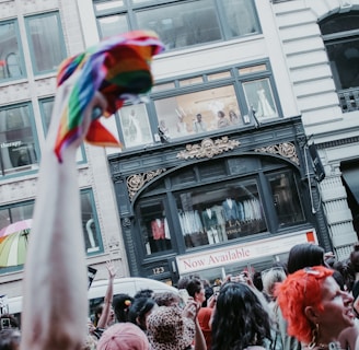 A vibrant crowd gathers in front of a city building with people looking out from a window above. There are diverse individuals, some holding rainbow flags, suggesting a celebratory or activist event. The architecture of the building features ornate decorations, and a sign reads 'Now Available.'