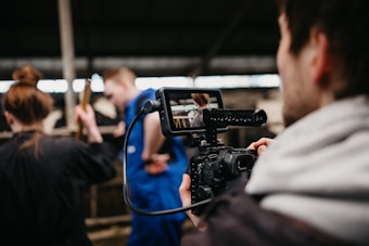 A person is filming with a professional camera, focusing on two individuals in work attire inside what seems to be an indoor farm setting. The individuals appear to be engaged in a task involving animals, possibly cows in the background.