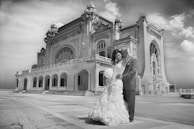 A couple dressed in wedding attire stands lovingly in front of an ornate, historic building with large arched windows and decorative architectural details. The sky is partly cloudy, captured in black and white.