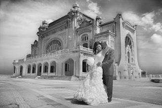A couple dressed in wedding attire stands lovingly in front of an ornate, historic building with large arched windows and decorative architectural details. The sky is partly cloudy, captured in black and white.