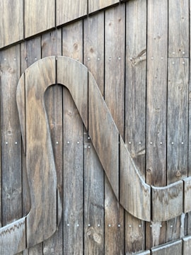 A wooden surface composed of vertical planks held together with small metal screws. A large stylized wooden letter or shape is attached to the planks, integrating seamlessly with the natural grain of the wood.