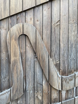 A wooden surface composed of vertical planks held together with small metal screws. A large stylized wooden letter or shape is attached to the planks, integrating seamlessly with the natural grain of the wood.
