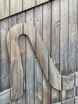 A wooden surface composed of vertical planks held together with small metal screws. A large stylized wooden letter or shape is attached to the planks, integrating seamlessly with the natural grain of the wood.