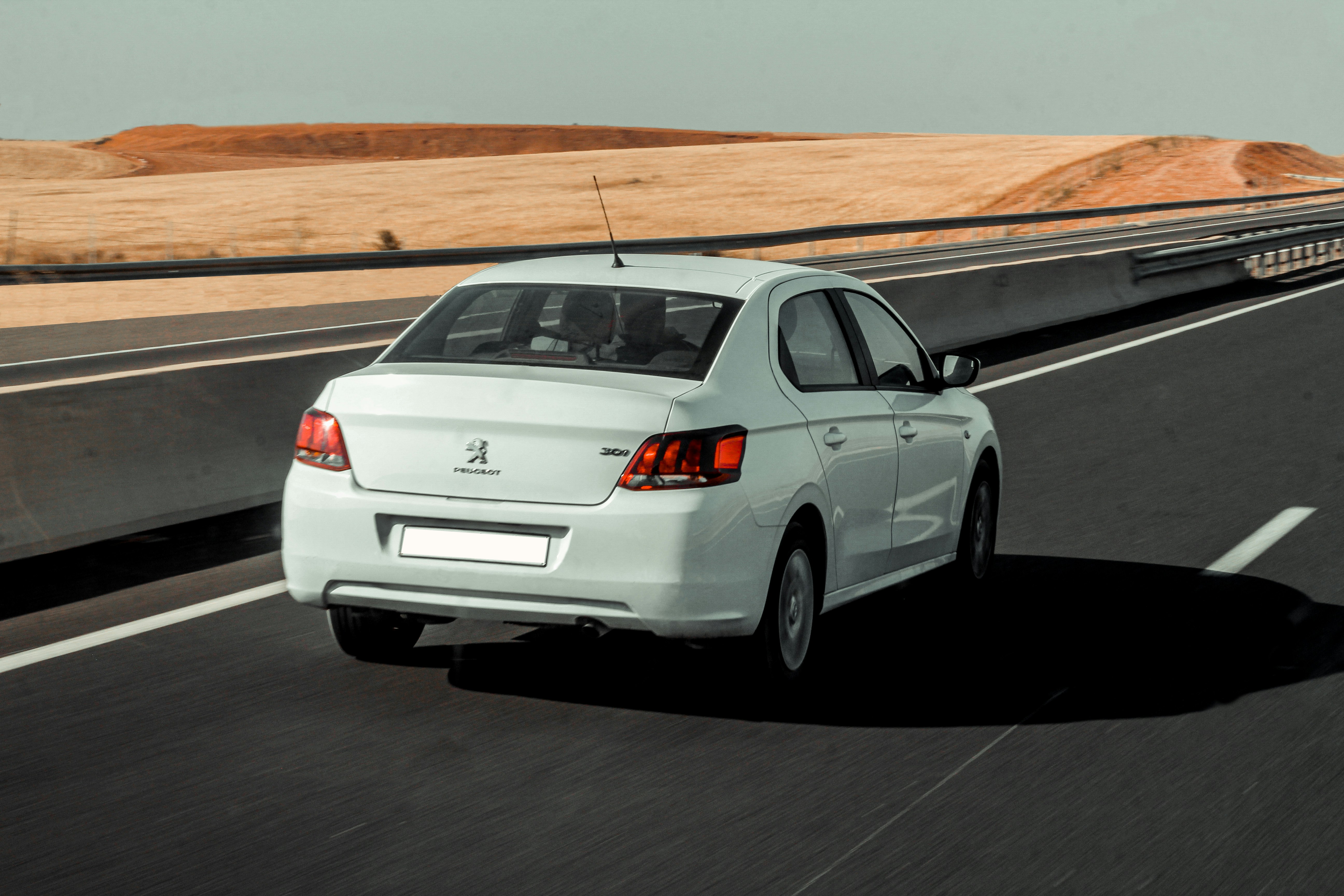 A white sedan drives along a highway past dry hills.