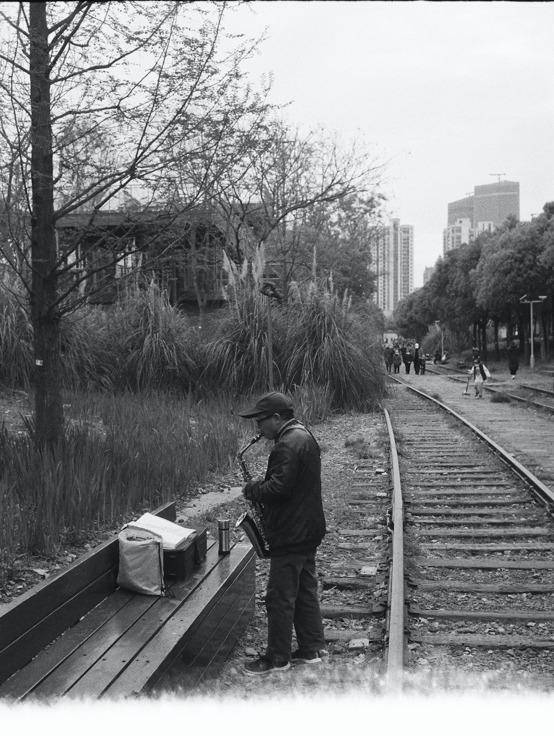 grayscale photo of man in jacket and pants holding umbrella walking on train rail