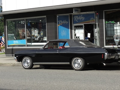 A black classic car with a convertible top, parked in front of a shop with large windows. The shop's signage includes the word 'Reckless' and has a blue color theme. The car has shiny chrome wheels and red interior seats.