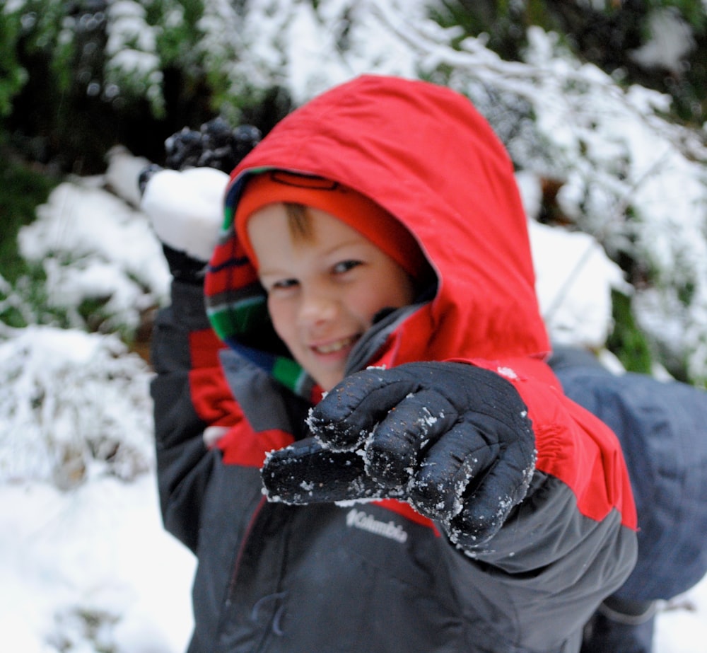 ragazzo in giacca con cappuccio nera e rossa su terreno coperto di neve durante il giorno