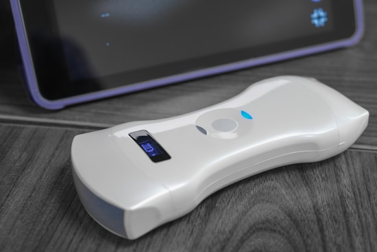 A white, handheld electronic device rests on a wooden surface. The device features a small digital display indicating battery status. Behind it, a section of a dark screen with a border is visible, suggesting the presence of another electronic device, possibly a tablet.