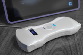 A white, handheld electronic device rests on a wooden surface. The device features a small digital display indicating battery status. Behind it, a section of a dark screen with a border is visible, suggesting the presence of another electronic device, possibly a tablet.