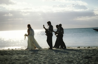 A bride in a white gown walks along a sandy beach followed by a group of musicians playing string instruments. The ocean gleams in the background under a partly cloudy sky.