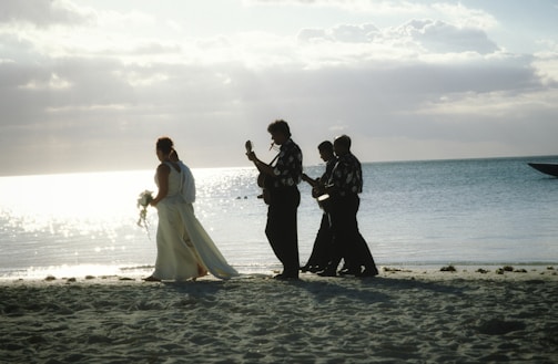 A bride in a white gown walks along a sandy beach followed by a group of musicians playing string instruments. The ocean gleams in the background under a partly cloudy sky.