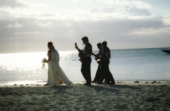 A bride in a white gown walks along a sandy beach followed by a group of musicians playing string instruments. The ocean gleams in the background under a partly cloudy sky.