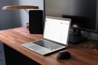 A modern workspace featuring a wooden desk with a laptop on it. The laptop is open, displaying a webpage in a well-lit room. A computer monitor is positioned behind the laptop, alongside a lamp. A mouse and a small coaster are also visible on the desk.