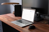 A modern workspace featuring a wooden desk with a laptop on it. The laptop is open, displaying a webpage in a well-lit room. A computer monitor is positioned behind the laptop, alongside a lamp. A mouse and a small coaster are also visible on the desk.