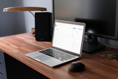 A modern workspace featuring a wooden desk with a laptop on it. The laptop is open, displaying a webpage in a well-lit room. A computer monitor is positioned behind the laptop, alongside a lamp. A mouse and a small coaster are also visible on the desk.