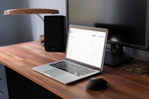 A modern workspace featuring a wooden desk with a laptop on it. The laptop is open, displaying a webpage in a well-lit room. A computer monitor is positioned behind the laptop, alongside a lamp. A mouse and a small coaster are also visible on the desk.