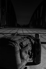 A black and white image displays a bridge casting long shadows across a concrete surface. In the foreground, a backpack and a stainless steel water bottle are placed, with the bridge structure receding into the distance.
