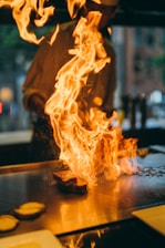 A chef stands in front of a grill with a tall flame rising dramatically as a piece of meat cooks. The background is slightly blurred, suggesting a bustling kitchen environment. The vivid fire creates an intense and dynamic scene.