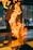 A chef stands in front of a grill with a tall flame rising dramatically as a piece of meat cooks. The background is slightly blurred, suggesting a bustling kitchen environment. The vivid fire creates an intense and dynamic scene.