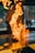 A chef stands in front of a grill with a tall flame rising dramatically as a piece of meat cooks. The background is slightly blurred, suggesting a bustling kitchen environment. The vivid fire creates an intense and dynamic scene.
