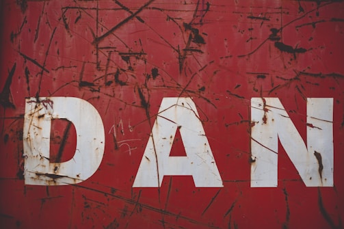 Large, bold white letters spelling 'DAN' are displayed on a weathered and rusty red metal surface. The surface is marked with scratches and patches of rust, suggesting age and exposure to the elements.
