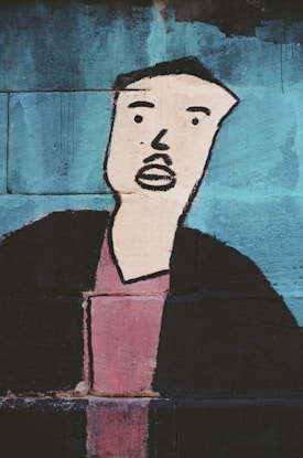 A graffiti-style painting of a person on a brick wall, featuring an abstract and minimalist depiction of a face with prominent black lines for facial features. The background is predominantly blue with the figure's shirt painted in a muted red and the jacket in black.