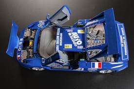 A detailed model of a racing car with its doors and parts of the engine compartment open, predominantly in blue with various decals and sponsor names. The model includes details like a roll cage, racing numbers, and national flags.