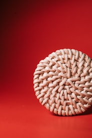 A close-up view of a round, woven basketry object placed on a vibrant red background. The texture of the weaving is prominent, highlighting the intricate craftsmanship and natural beige color of the material.