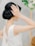 A woman in a white textured sleeveless gown with an elegant updo hairstyle. Her hand is raised towards her head, touching her hair gently. In the blurred background, there are green and white floral decorations.