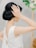 A woman in a white textured sleeveless gown with an elegant updo hairstyle. Her hand is raised towards her head, touching her hair gently. In the blurred background, there are green and white floral decorations.