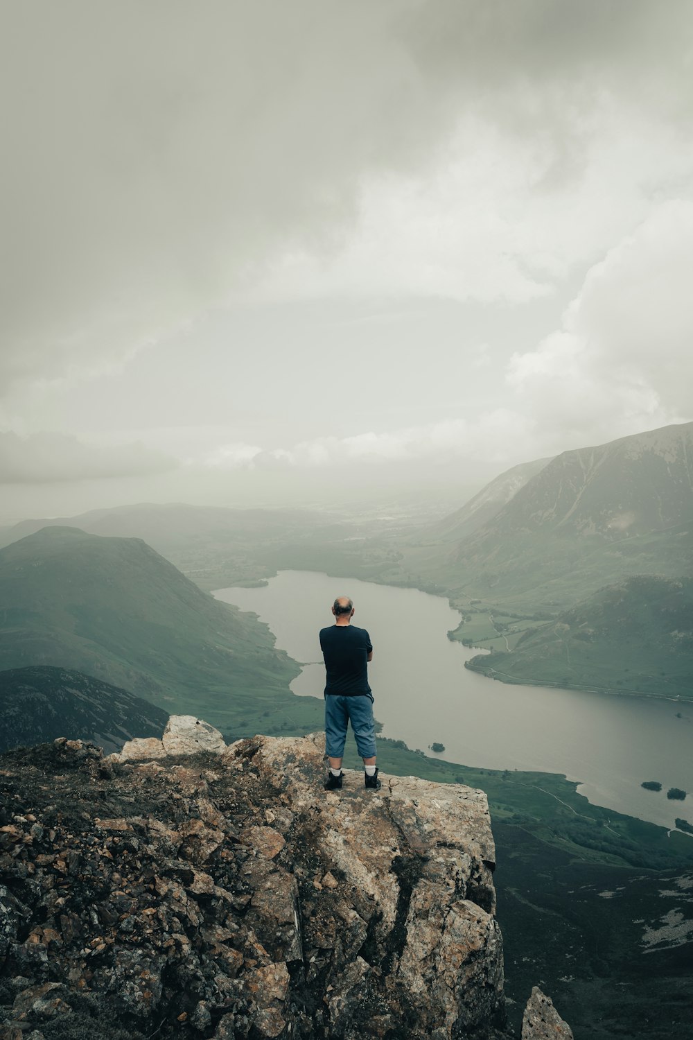 man in black jacket standing on rock formation looking at lake during daytime